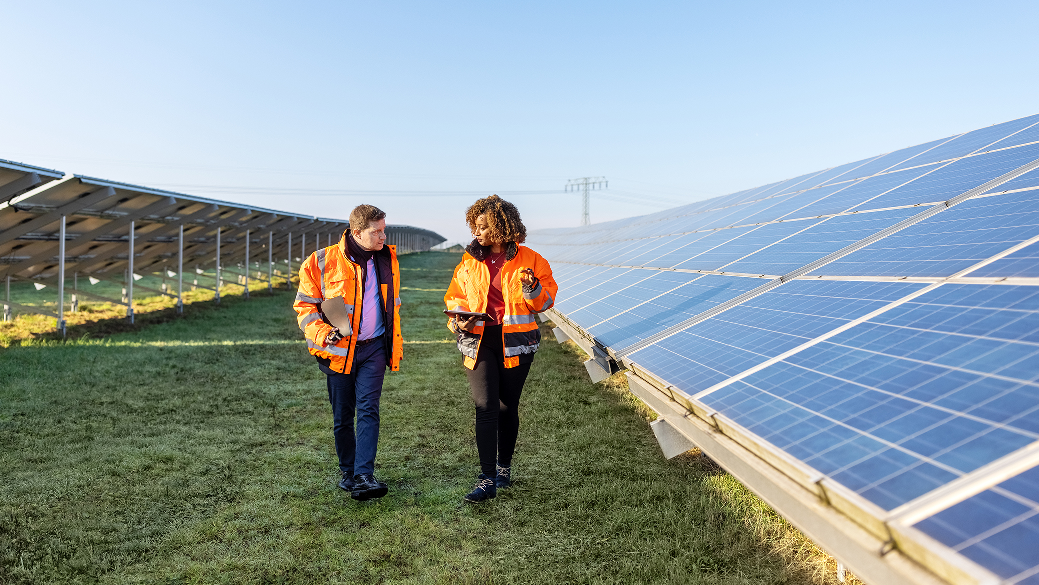 Male and female engineers working at solar power plant. Two technicians in reflective clothing walking between rows of photovoltaic panels at solar farm.